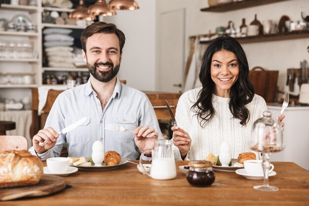 Portrait of happy couple man and woman eating at table while having breakfast in kitchen at home