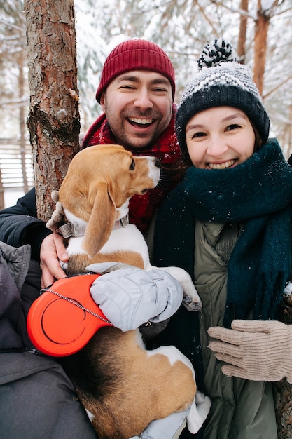 Portrait of a happy couple standing outdoors at winter with their beagle dog. Both wearing hats, jackets and scarfs. Conifer tree trunks in background.