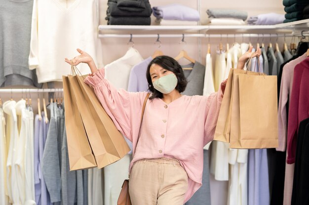 衣料品店に立っている彼女の手で購入品を保持している保護マスクで幸せな顧客の肖像画