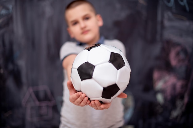 portrait of happy cute boy having fun holding a soccer ball while standing in front of black chalkboard