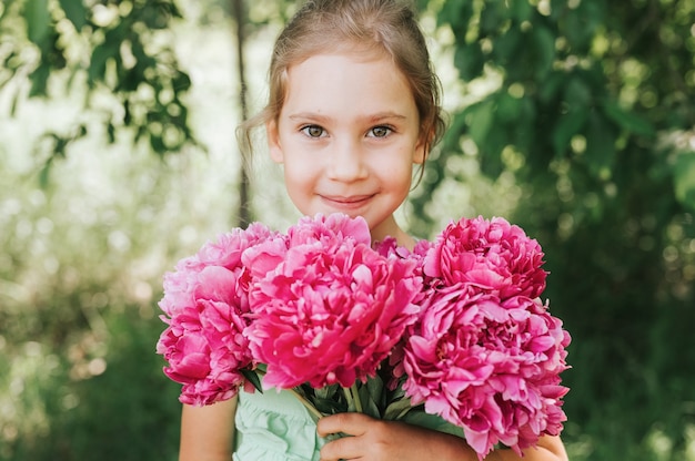 Portrait of a happy cute little caucasian seven year old kid girl, holds in hands a bouquet of pink peony flowers in full bloom on the green background of nature