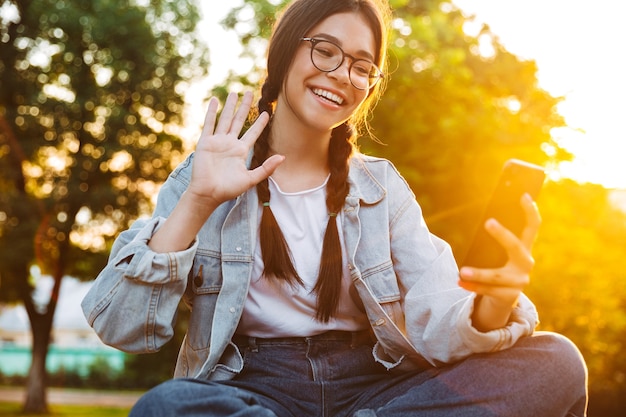 Portrait of a happy cute young student girl wearing eyeglasses sitting on bench outdoors in nature park with beautiful sunlight talking by mobile phone waving