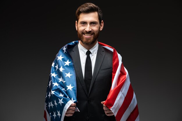 Portrait of happy dark-haired young man in business style\
clothes wrapped in flag of america and smiling to camera.\
citizenship in america, immigration concept