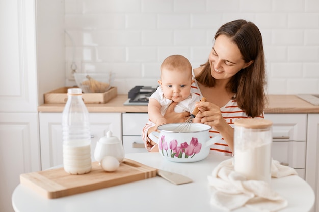 Portrait of happy delighted woman with dark hair sitting at table with infant baby daughter mommy holding whisk together with kid cooking bakingin kitchen