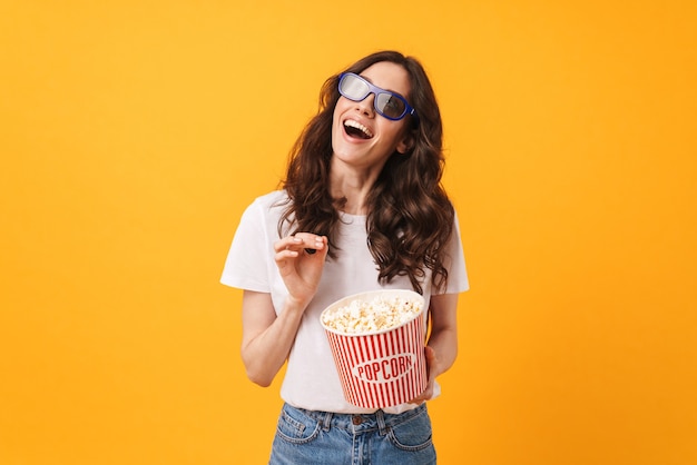 Portrait of happy dreaming young woman posing isolated over yellow wall eat popcorn watch film.