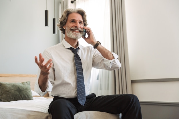 Портрет счастливого эмоционального старшего седого делового человека, сидящего на кровати в помещении дома, разговаривая по мобильному телефону.