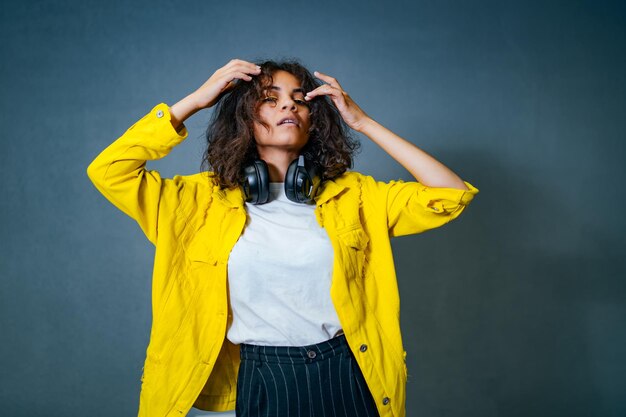 Portrait of happy emotional teenage girl with headphones feeling confident while posing to the camera. wearing bright yellow cotton jacket and white shirt. grey wall background