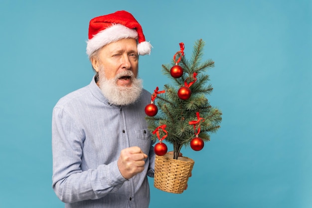 Portrait of happy emotions santa claus excited looking at camera and holds small christmas tree on b