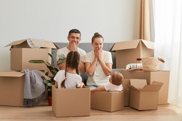 Portrait of happy excited family, woman and man with their children sitting on floor with excited surprised facial expression, kids in cardboard boxes, relocating in a new house.