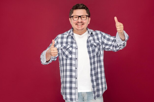 Portrait of happy excited middle aged business man in casual checkered shirt and eyeglasses standing, thumbs up, toothy smiling looking at camera. indoor studio shot, isolated on dark red background.