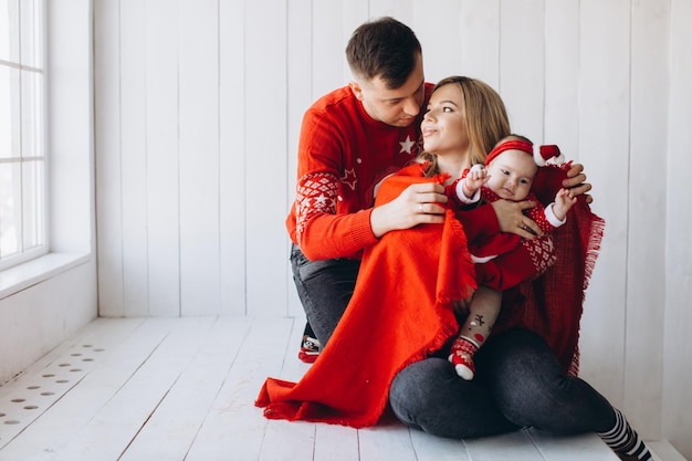 Portrait of happy family mom dad and little daughter in red traditional christmas clothes spending time together in light wooden room near window copy space