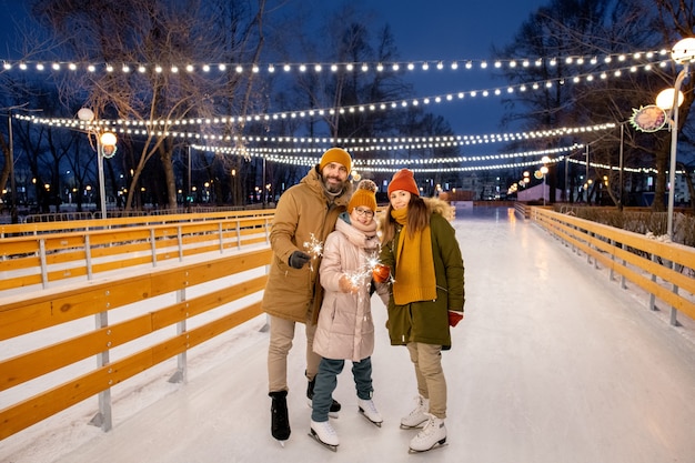 Ritratto di una famiglia felice di tre persone con stelle filanti in piedi sulla pista di pattinaggio all'aperto durante le vacanze di natale