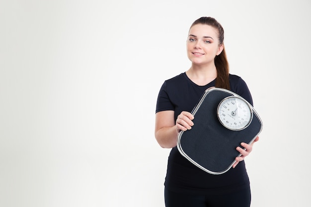 Portrait of a happy fat woman holding weighing machine isolated on a white wall