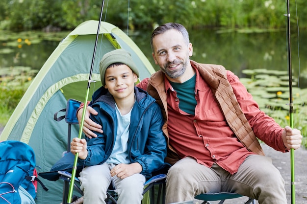 Portrait of happy father embracing son while enjoying fishing trip together and smiling at camera