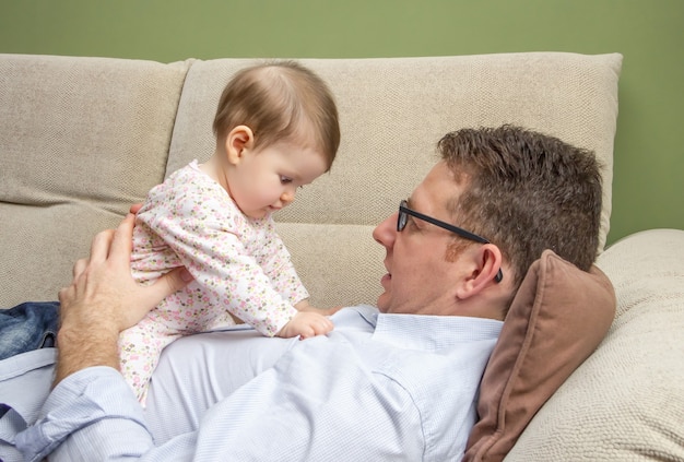 Portrait of happy father playing with cute baby sitting over his belly in a sofa at home