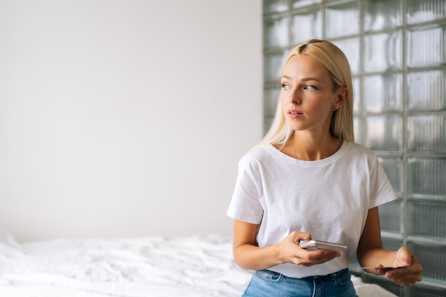 Portrait of happy female shopper thinking about sales holding credit card and smartphone sitting on bed in light living room pensive looking away