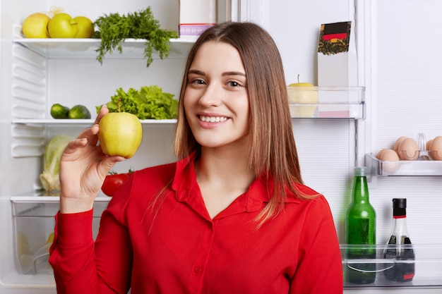 Portrait of happy female vegeterian holds fresh apple, stands near fridge with happy expression, keeps to diet, has fridge full of vegetables