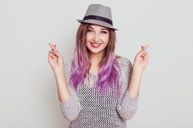 Portrait of happy female with pleasant appearance, crossing fingers, hopes for better, looking smiling at camera, wishes dream comes true, isolated over grey background.