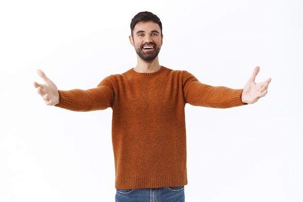 Portrait of happy and friendly bearded man give warm welcome to dear guest, spread hands sideways in cuddle or embraces, smiling greeting person with love, standing white wall