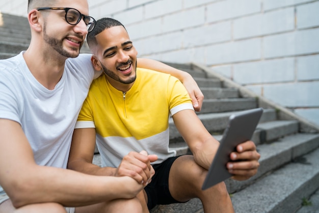 Portrait of happy gay couple spending time together and taking a selfie with digital tablet while sitting on stairs. Lgbt and love concept.