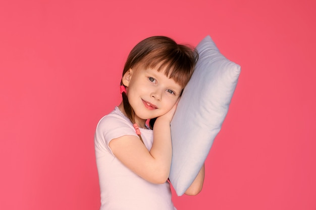 Portrait of a happy girl 5-6 years old on a pink wall with a pillow in her hands, the child is preparing for bed.