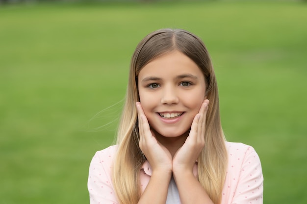 Portrait of happy girl child smiling with hands around face on green grass background outdoors