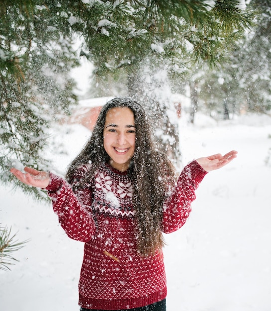 Portrait of a happy girl in a christmas sweater in the winter forest snow falls from a pine branch