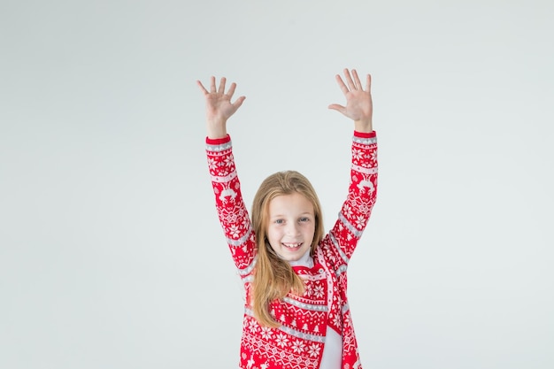 흰색 배경에 격리된 크리스마스 스웨터를 입고 머리 위로 손을 들고 춤추는 행복한 소녀의 초상화크리스마스와 새해 개념빨간 스웨터를 입은 긍정적인 소녀크리스마스 분위기