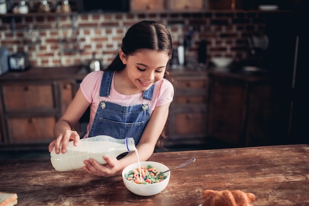 Photo portrait of happy girl pouring milk into the bowl of colorful corn flakes for breakfast in the kitchen