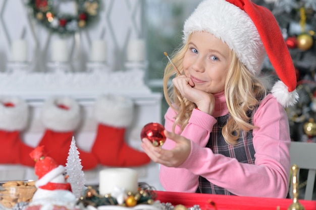 Portrait of happy girl in Santa hat prapring for  Christmas sitting at the table and holding Christmas decoration