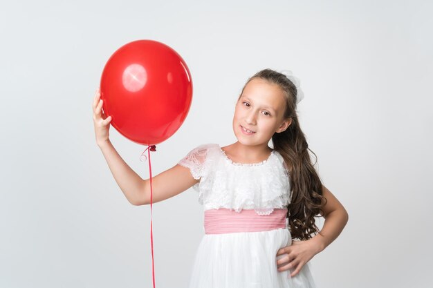 Portrait of happy girl in white dress holding one red balloon in hand smiling and looking at camera