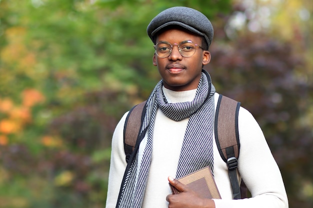 Portrait of happy handsome black African college or university student, Afro American young intelligent smart man reader with backpack and book in hand in scarf, glasses and hat walking in autumn park