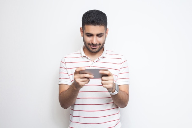 Portrait of happy handsome blogger young adult man wearing T-shirt standing, using smartphone and texting message with smile, looking video. Indoor, isolated, studio shot, white background