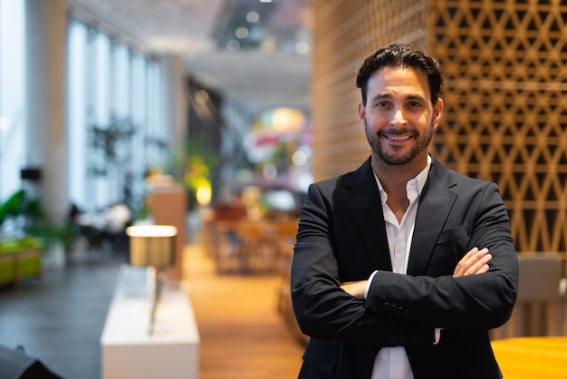 Portrait of happy handsome Hispanic businessman at coffee shop with arms crossed