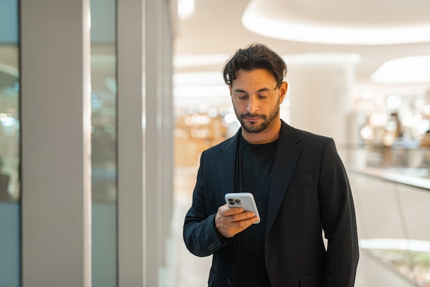 Ritratto di felice uomo d'affari ispanico bello utilizzando il telefono accanto alla finestra al centro commerciale