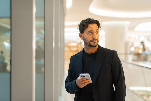 Ritratto di felice uomo d'affari ispanico bello utilizzando il telefono accanto alla finestra al centro commerciale