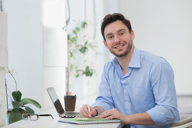 Portrait of happy handsome male freelancer working in restautant with laptop computer. Cute man in blue shirt writing something in notebook.