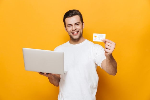 Portrait of a happy handsome man wearing t-shirt standing isolated over yellow wall, showing credit card