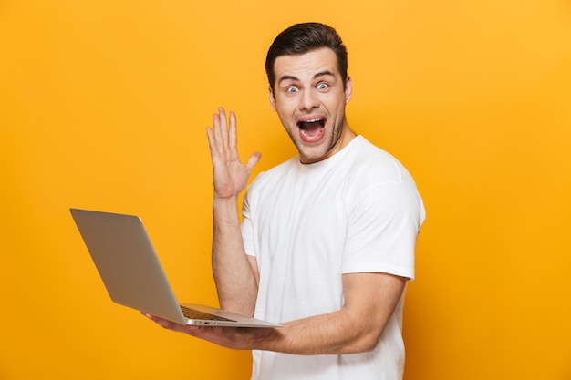 Portrait of a happy handsome man wearing t-shirt standing isolated over yellow wall, using laptop computer, celebrating