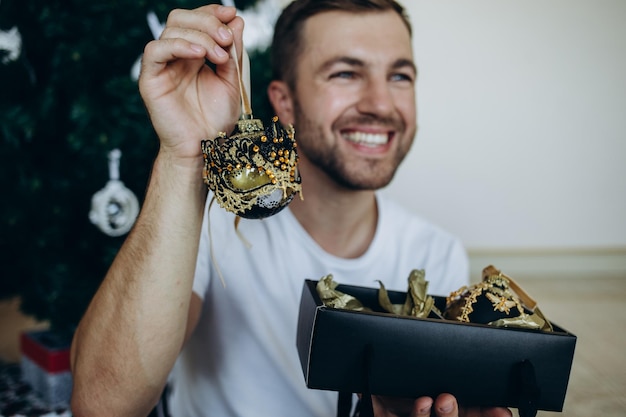 Portrait of happy handsome young man holding cardboard box with green ribbon near Christmas tree and looking at camera