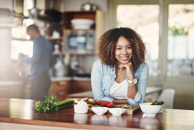 Portrait of a happy healthy and carefree young woman preparing a healthy meal at home with her husband in the background Black wife making an organic vegetarian salad for lunch in a kitchen