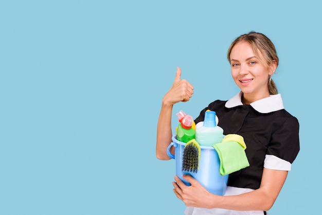 Portrait of happy housekeeper showing thumb up gesture holding cleaning equipment's in bucket looking at camera