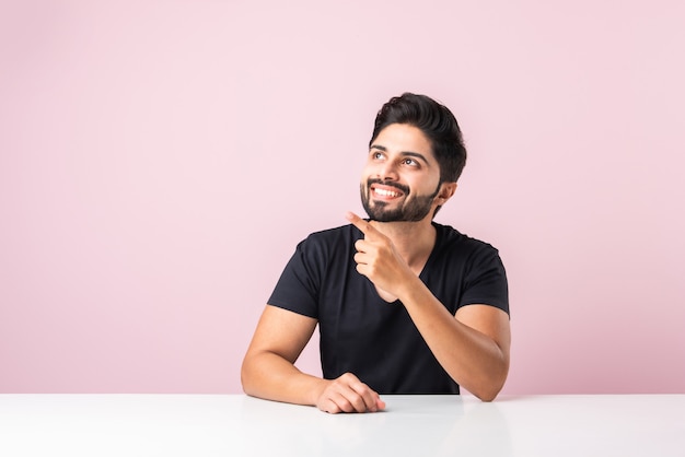 Portrait of happy indian asian bearded man sitting at table or\
desk or platform against pink background looking at camera