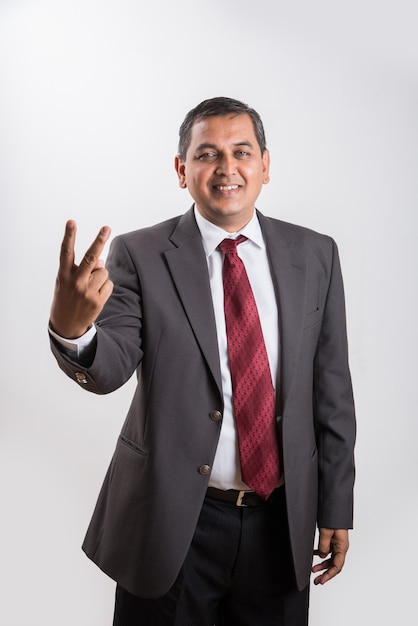 Portrait of happy Indian or Asian mid aged businessman in black suit with success sign while standing isolated over white background