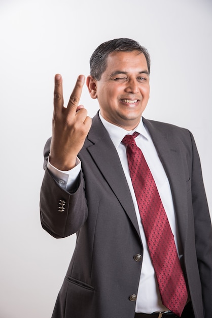 Portrait of happy Indian or Asian mid aged businessman in black suit with success sign while standing isolated over white background