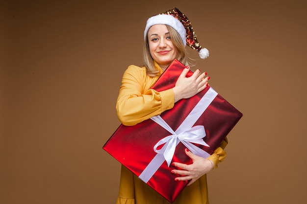 Portrait of happy and jovial blonde woman in sparkling Santa hat and yellow dress hugging wrapped red Christmas presents with her eyes closed