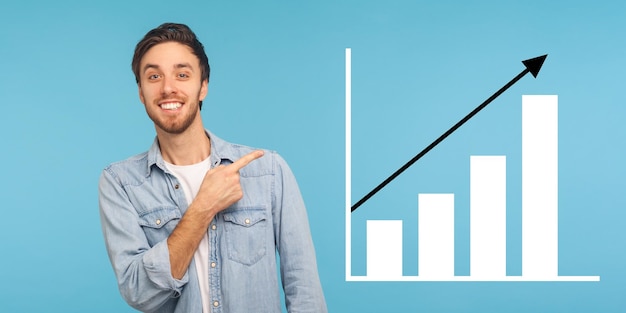 Портрет счастливого радостного молодого бородатого мужчины, стоящего в стороне и показывающего график роста бизнеса в помещении студии, снятой на синем фоне