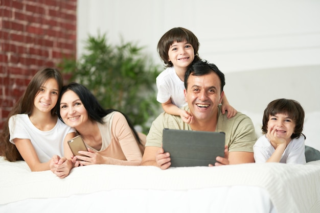 Portrait of happy latin family with cute kids smiling at camera while using digital devices, lying on the bed together. Family, parenthood, technology concept