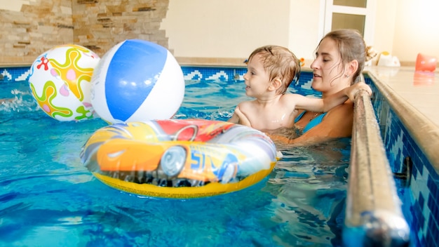 Portrait of happy laughing toddler boy with young mother playing with colorful inflatable beach ball in swimming pool at summer hotel resort