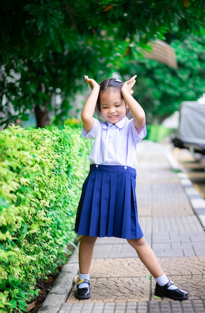 学校に戻って準備ができて、公園に立っているタイの学校の制服で幸せな小さなアジアの女の子の肖像画